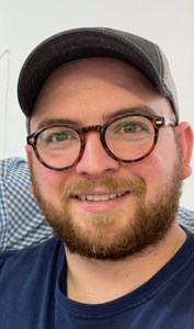 man with classes and beard smiling and wearing a ball cap.